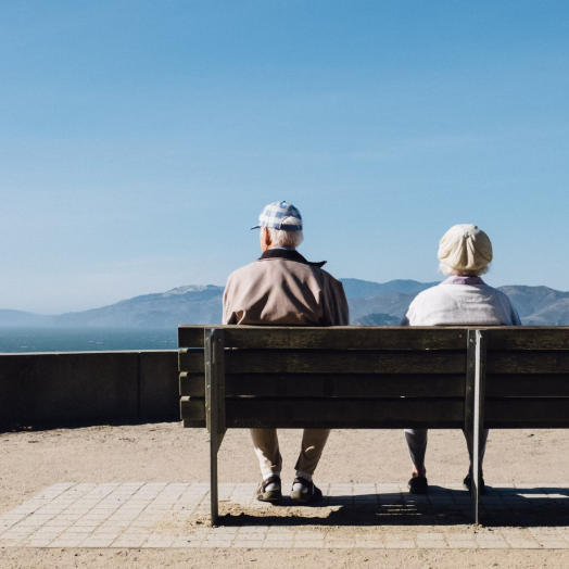 A mature couple sitting on a park bench staring out at a lake and mountains.